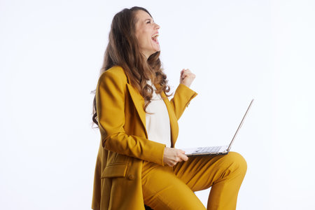 An empowered woman in a yellow business suit triumphantly celebrates success with a fist pump while holding her laptop, conveying determination and achievement.の写真素材