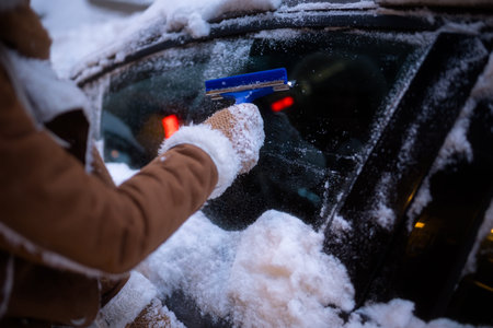 A person in warm clothing uses a blue tool to scrape ice and snow off a car windshield at night, with city lights blurred in the background.の写真素材