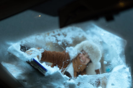 A young woman in a fur hat smiles while using an ice scraper to clear the windshield from inside the car, preparing for a winter drive.の写真素材