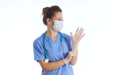 A professional female healthcare worker in blue scrubs and a face mask puts on sterile latex gloves,...
