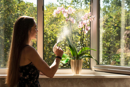 A woman waters the orchid flowers in front of the opened window