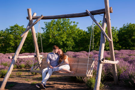 Lovely couple sits on a rustic wooden swing amidst lavender field on a sunny summer day. The man embraces the woman, who leans on his shoulder, creating a serene and romantic sceneの写真素材