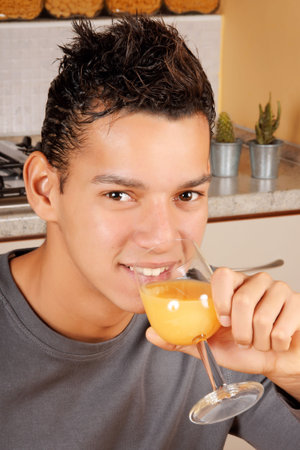 Young brazilian man drinking a glass of orange juice in the kitchen at homeの写真素材