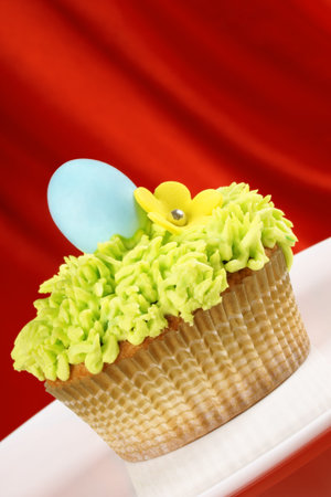 Fancy Easter cupcake decorated with sugar flower and hard sugar coated chocolate egg over a red...
