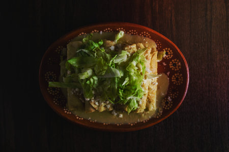 Green Enchiladas with lettuce served in a clay dish on a wooden table.