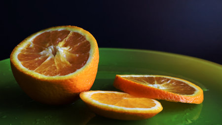 Half an orange and slices on a green glass plate with drops of water, on a table close-up, on a black background. The fruit is multi-nested, multi-seeded, with a thick two-layer peelの写真素材