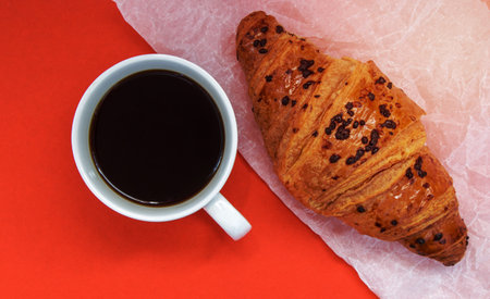 Black coffee without milk in a white cup and a chocolate croissant on parchment and bright background. French breakfast with fresh pastries. Top view flat with copy space for your textの写真素材