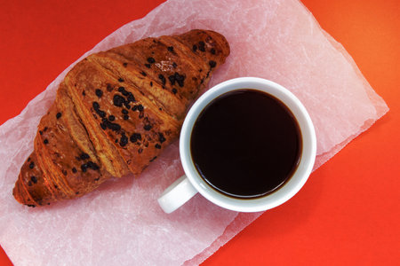 Black coffee without milk in a white cup and a chocolate croissant on parchment and bright background. French breakfast with fresh pastries. Top view flat with copy space for your textの写真素材