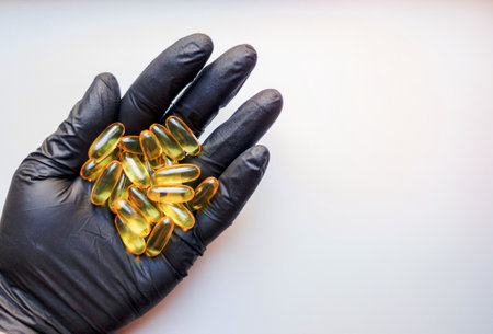 fish oil capsules in hand in a black medical glove on a white background, the hand takes one tablet from a saucer or plate. Hand holding omega 3 capsule on a white background. Healthcare concept.の写真素材
