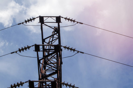 High voltage electrical tower. Power lines and power pylons on a sunny day with circular clouds in the blue sky. High voltage power transmission line supports. They have a complex steel structureの写真素材