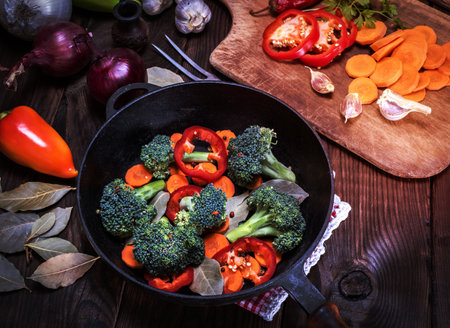 fresh pieces of broccoli, carrots and red peppers in a black cast-iron frying pan on a brown table, top viewの写真素材
