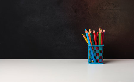 metal glass with pens, pencils and felt-tip pens and scissors on the background of an empty black chalk board, white table, copy spaceの写真素材
