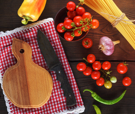 empty wooden cutting board with a knife and fresh red cherry tomatoes, beside a tied bundle of long raw spaghettiの写真素材