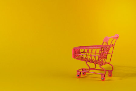 empty pink miniature shopping cart on yellow background. Seasonal sale, copy spaceの写真素材