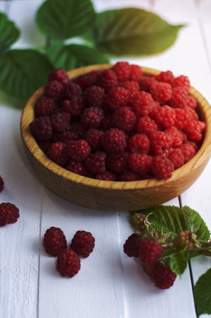 Fresh raspberries in a wooden bowl with green leaves from a bush on a white tableの写真素材