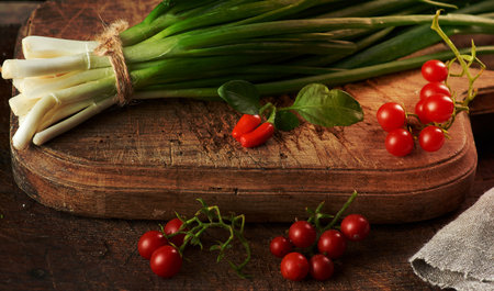 red ripe cherry tomatoes and a bundle of green onions tied with a rope lies on a brown cutting boardの写真素材