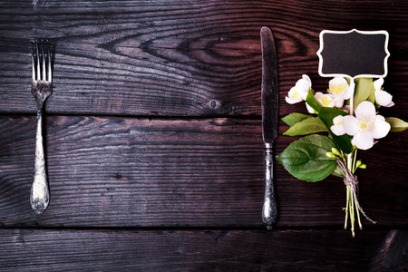 Iron fork and knife with a bouquet of jasmine on a brown wooden background, empty space in the middleの写真素材
