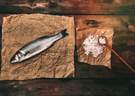 fresh whole sea bass fish on brown crumpled paper and salt with a wooden spoon, top view, copy spaceの写真素材