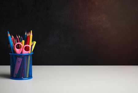 metal glass with pens, pencils and felt-tip pens and scissors on the background of an empty black chalk board, white table, copy spaceの写真素材