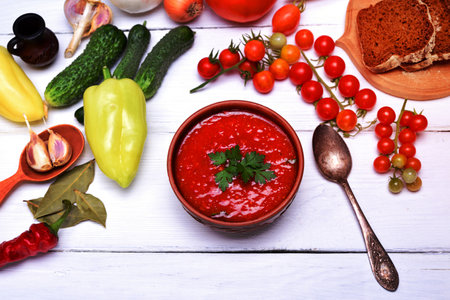 Soup gazpacho in a brown ceramic plate on a white wooden background, top view, around fresh vegetables and herbs for cookingの写真素材