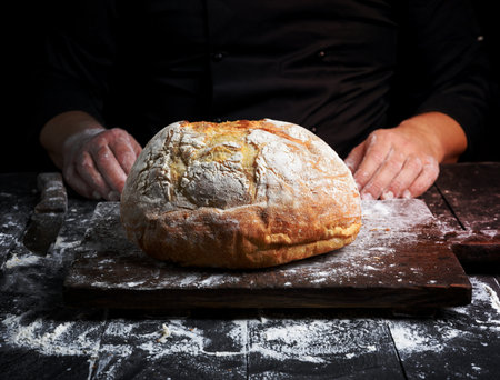 round baked homemade bread on an old brown wooden board and chef handsの写真素材