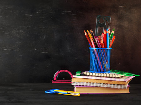 stack of notebooks, blue glass with multi-colored wooden pencils and pens, rulers and an eraser on the background of an empty black chalk board, back to school conceptの写真素材