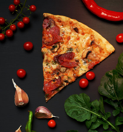 triangular slice of baked pizza with mushrooms, smoked sausages, tomatoes and cheese, next to fresh green leaves of arugula, black background, flat layの写真素材