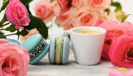 white ceramic cup with coffee and  macarons on a white table, behind a bouquet of pink roses, close upの写真素材