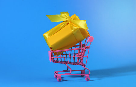 yellow gift box with a silk bow in a miniature metal trolley on a light blue background. Party Backdrop, Surprise, Seasonal Saleの写真素材