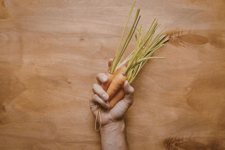 A hand holding a fresh carrot with green leaves on a wooden background