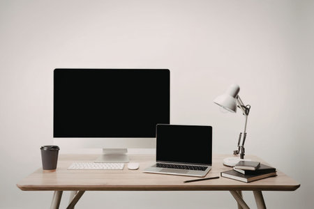 A modern workspace setup featuring a computer monitor, laptop, desk lamp, books, and a cup, all...