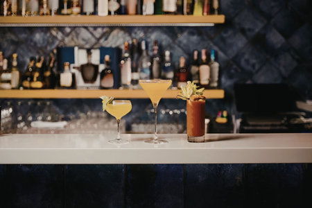 Two colorful cocktails on a bar counter with shelves of bottles in the background.