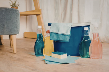 A collection of cleaning supplies on a wooden floor, including bottles, a bucket, and a sponge.