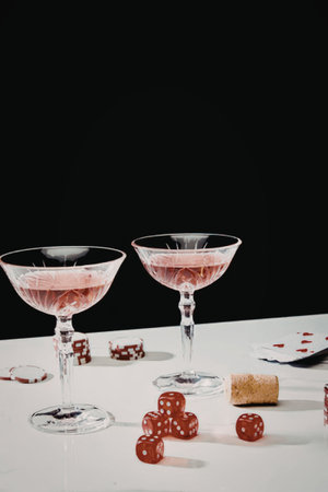 Two glasses of pink cocktail on a white table with red dice and a wine cork