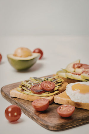A wooden cutting board with avocado toast, a fried egg, and cherry tomatoes on a white table.
