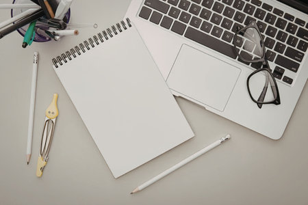 A laptop, spiral notebook, pens, pencils, glasses, and other office supplies on a desk.