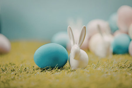 A small white Easter bunny sitting on green grass with a blue egg and other colorful eggs in the...