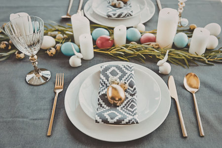 A beautifully set table with a theme, featuring a white plate with a gold ornament on a blue and white napkin, gold utensils, and a festive centerpiece with candles and ornaments.の写真素材