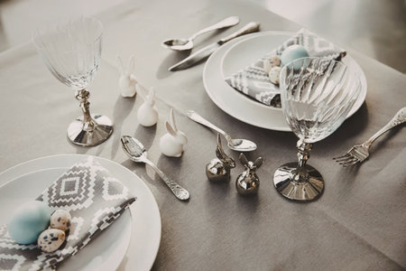 A beautifully set table with white plates, silverware, and wine glasses, adorned with blue Easter...