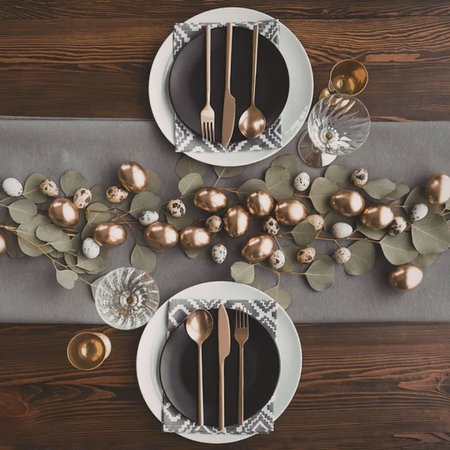 An overhead view of a table setting with two plates, each set with gold utensils, surrounded by...