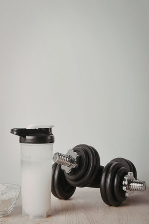 A clear plastic shaker bottle with a black lid containing a white liquid, likely protein shake, sits next to a pair of black dumbbells and a small glass bowl on a light-colored wooden surface.の写真素材
