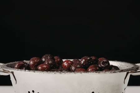A metal colander filled with fresh cherries on a white surface against a black background.の写真素材