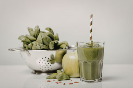 A glass of green smoothie with a straw, accompanied by a bowl of spinach, an apple, and chia seeds...