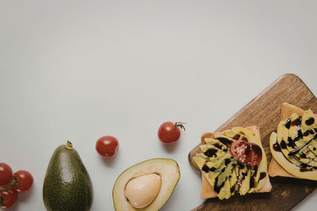 A wooden cutting board with two slices of avocado toast, cherry tomatoes, and a whole avocado on a...