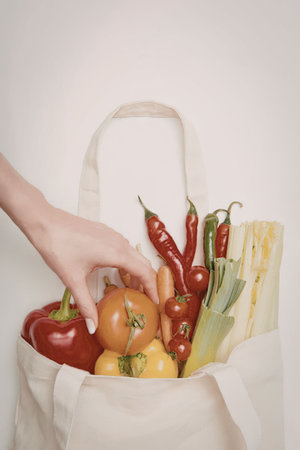 A hand reaching into a canvas bag filled with various fresh vegetables, including tomatoes, peppers, and celery.の写真素材