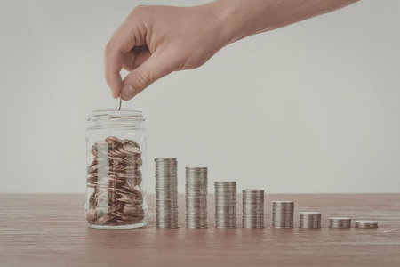 A hand putting a coin into a jar filled with coins, with additional coins stacked beside it,...