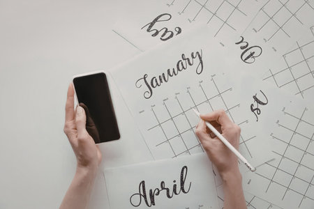 A person's hands are seen holding a phone and a pen, poised over a calendar with the months January...
