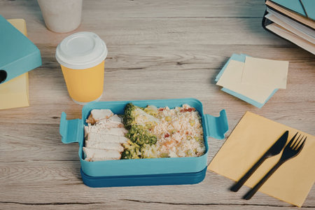 A blue lunch box with food, a cup of juice, and office supplies on a wooden table.