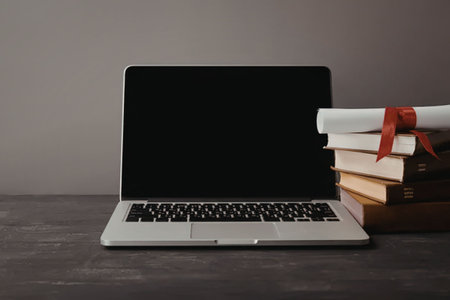 A silver laptop with a black screen and keyboard sits on a dark gray desk next to a stack of books tied with a red ribbon.の写真素材