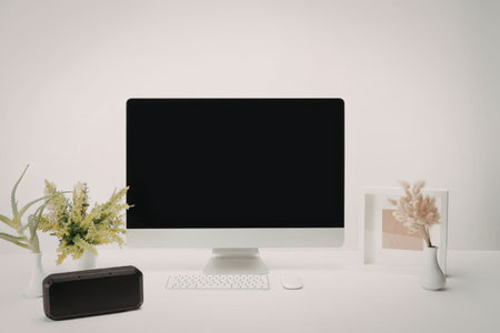 A modern workspace setup with a computer monitor, keyboard, speaker, plant, and vase on a white desk...